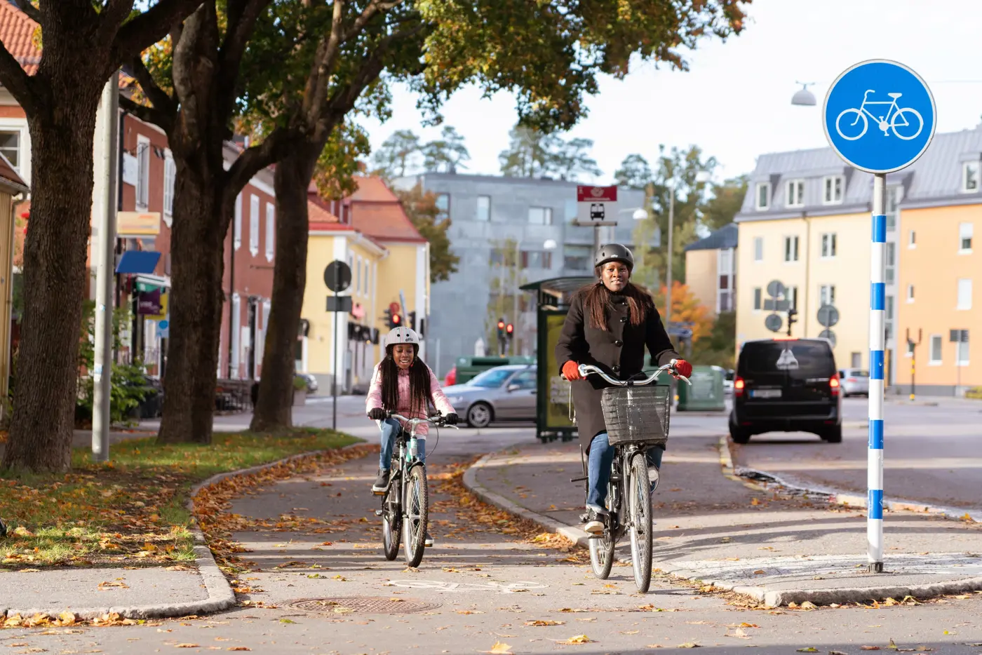 En mamma och ett barn cyklar bredvid varandra på en gång- och cykelbana i stadsmiljö.